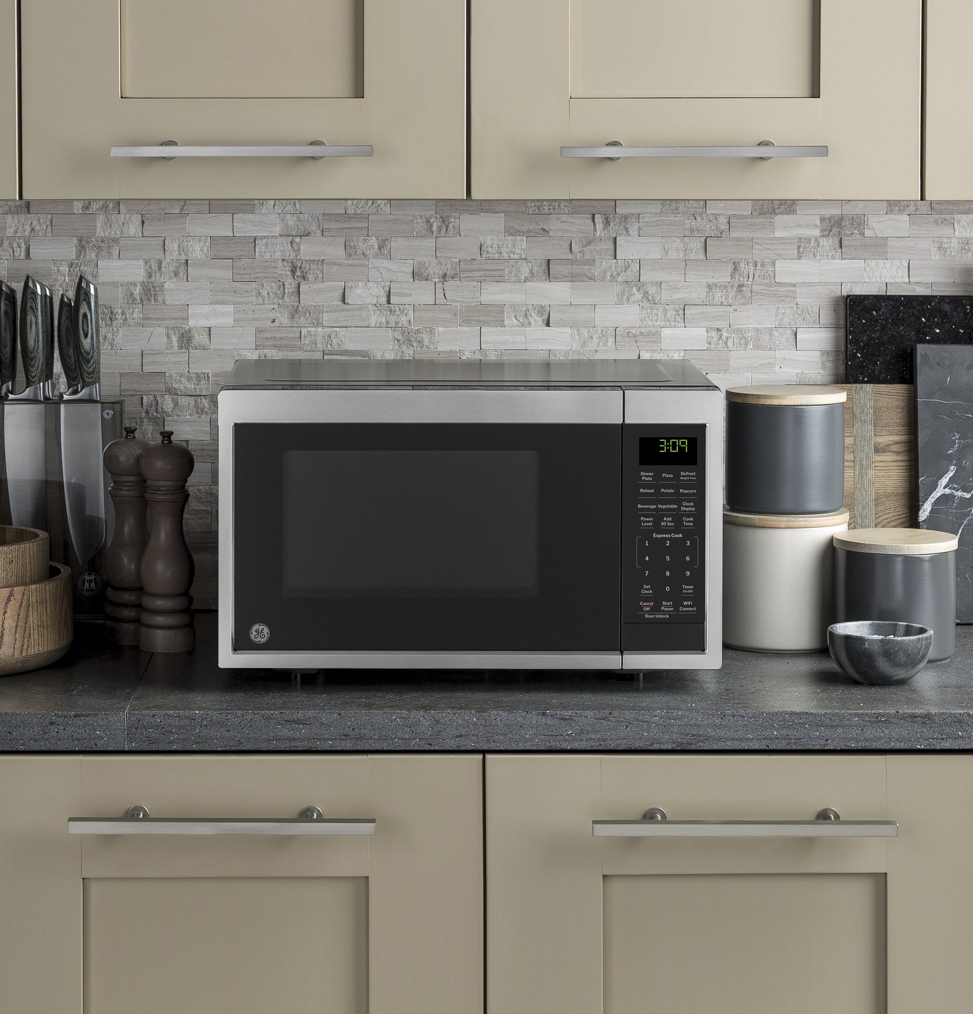 Stainless steel smart countertop microwave on kitchen counter with stone backsplash and storage jars nearby