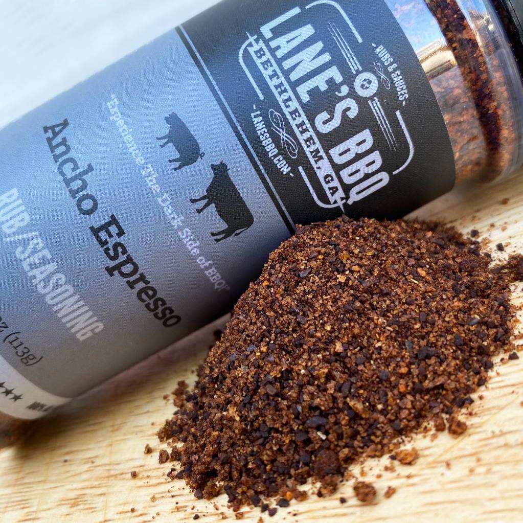 Dark coarse BBQ rub seasoning spilled from tilted black and gray Lane’s BBQ container on wooden surface
