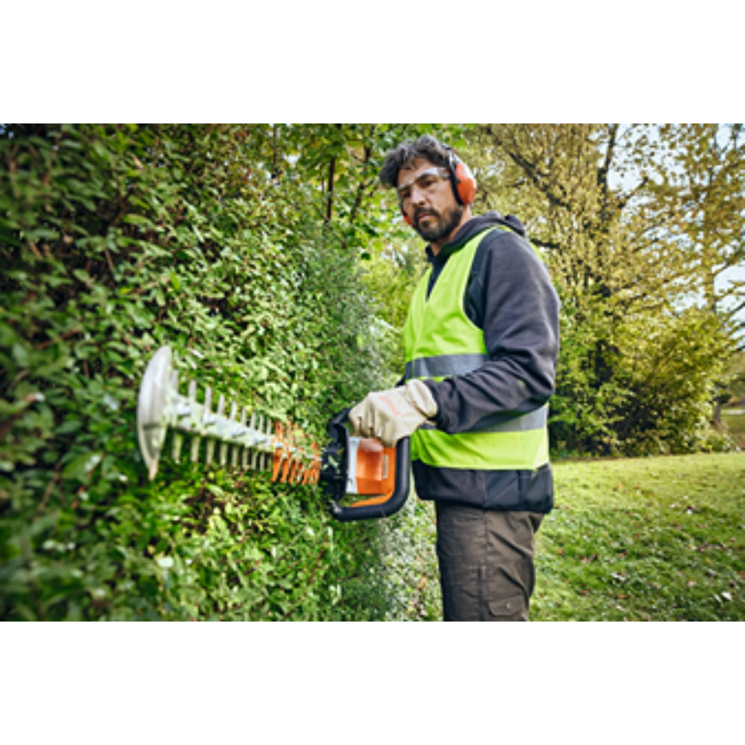 Man wearing safety gear using battery-powered hedge trimmer to shape a green hedge for home improvement tasks