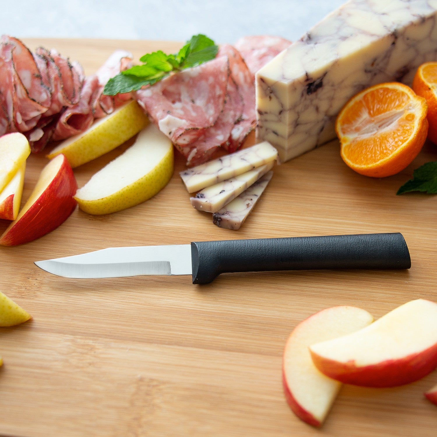 Peeling paring knife with black handle on wooden board surrounded by sliced fruit, cheese, and cured meat for kitchen prep