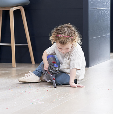 Young child using a Casdon cordless vacuum toy to clean confetti from a light wood floor in a modern room setting