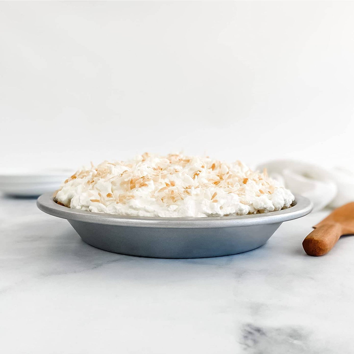 Round metal pie pan filled with whipped topping and toasted coconut flakes on a light marble surface