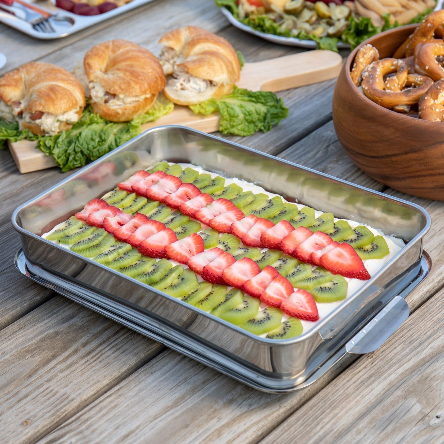 Rectangular stainless steel cake pan with lid removed, filled with dessert topped with sliced kiwi and strawberries on wooden table