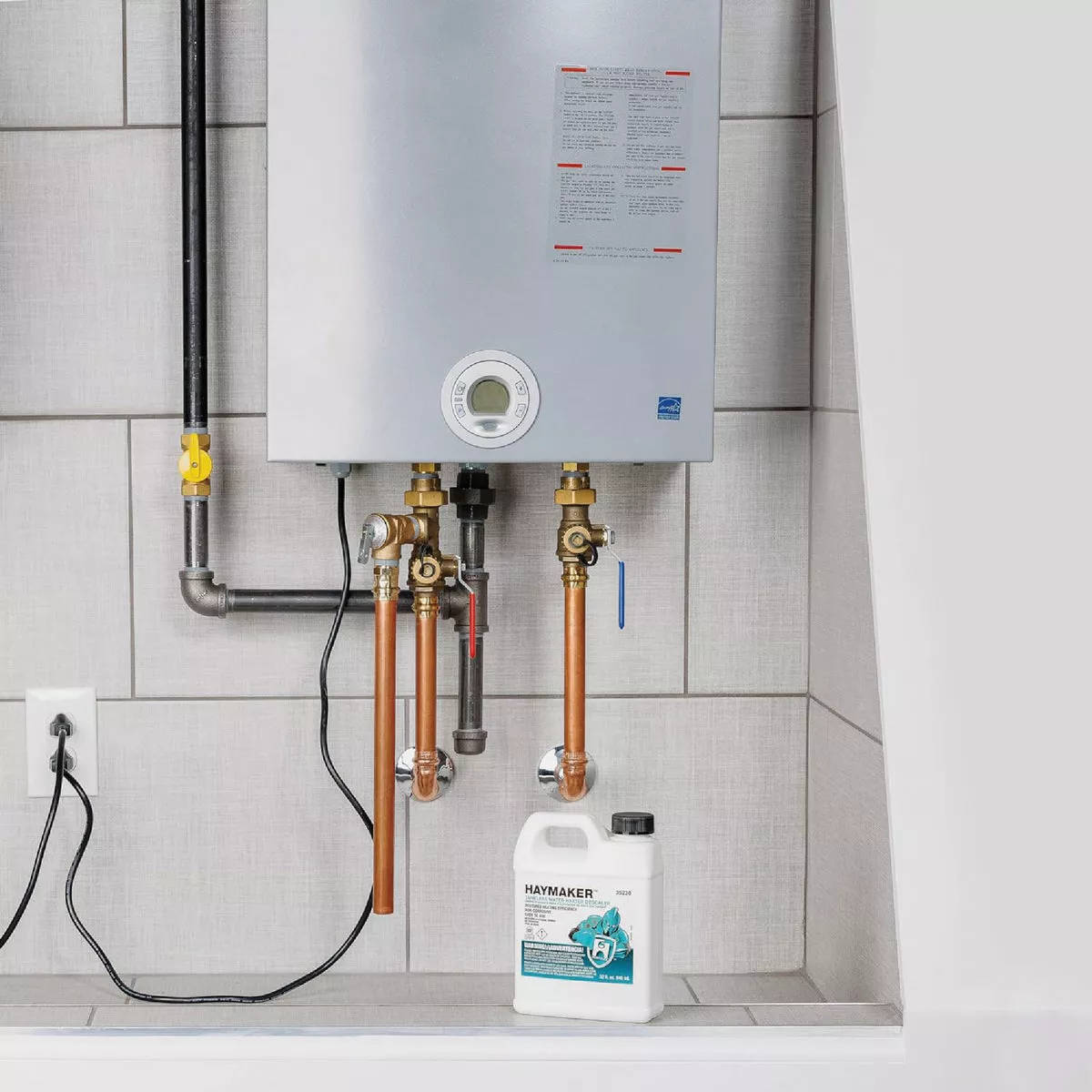 White container of Hercules Haymaker descaler placed below tankless water heater with copper pipes and control panel visible