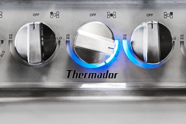 Close-up of stainless steel gas cooktop control knobs with blue burner flames in kitchen appliances collection