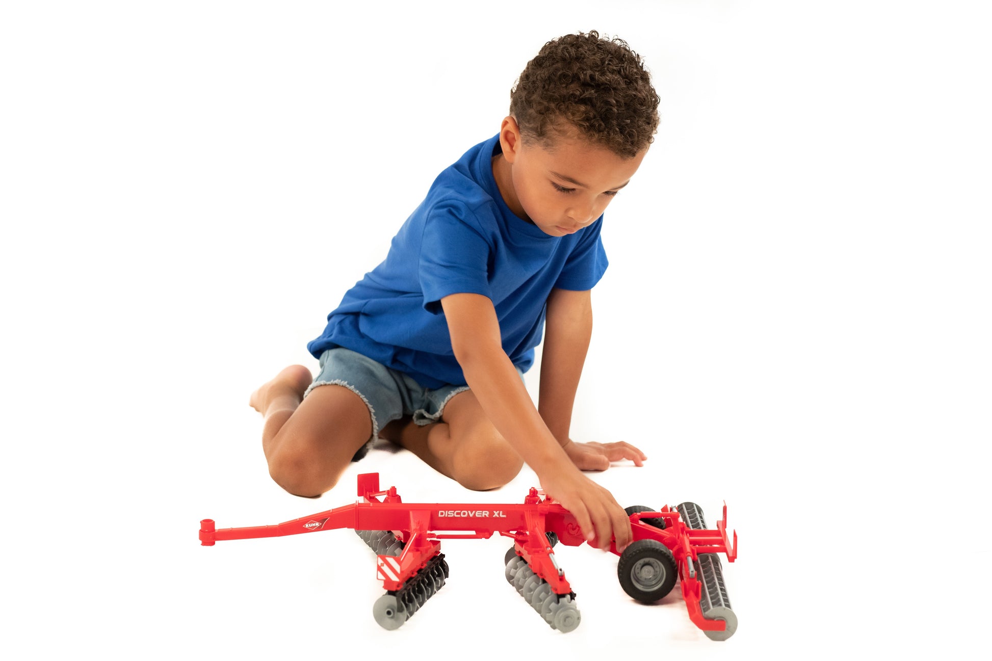 Child in blue shirt kneeling and engaging with a red Bruder Kuhn Disc Harrow Discover XL toy for creative play