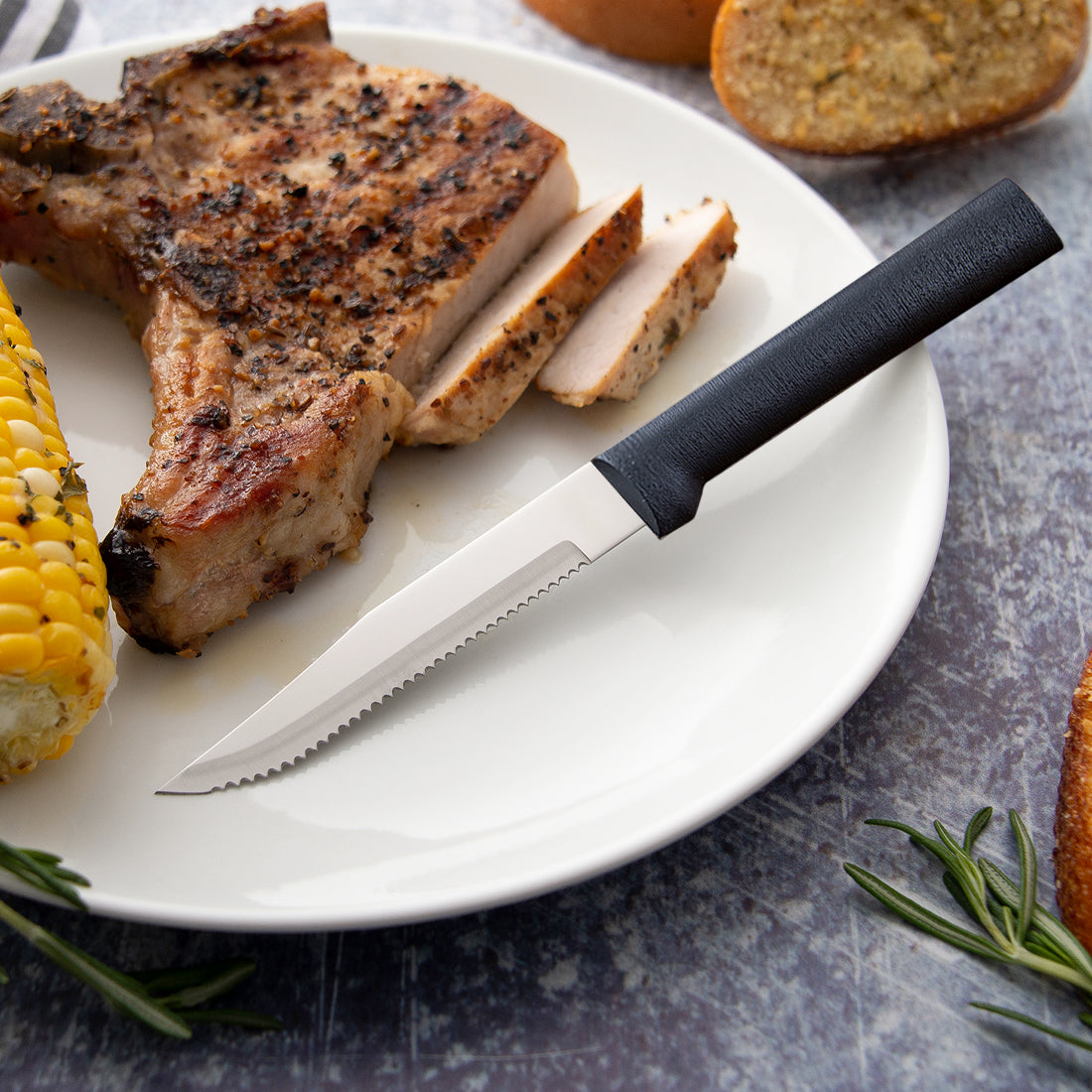 Serrated steak knife with black handle on white plate with grilled pork chop, corn, and bread in kitchenware collection under $50
