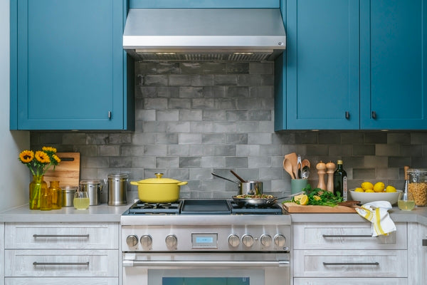 Stainless steel custom insert range hood above gas cooktop in modern kitchen with blue cabinets and stone backsplash