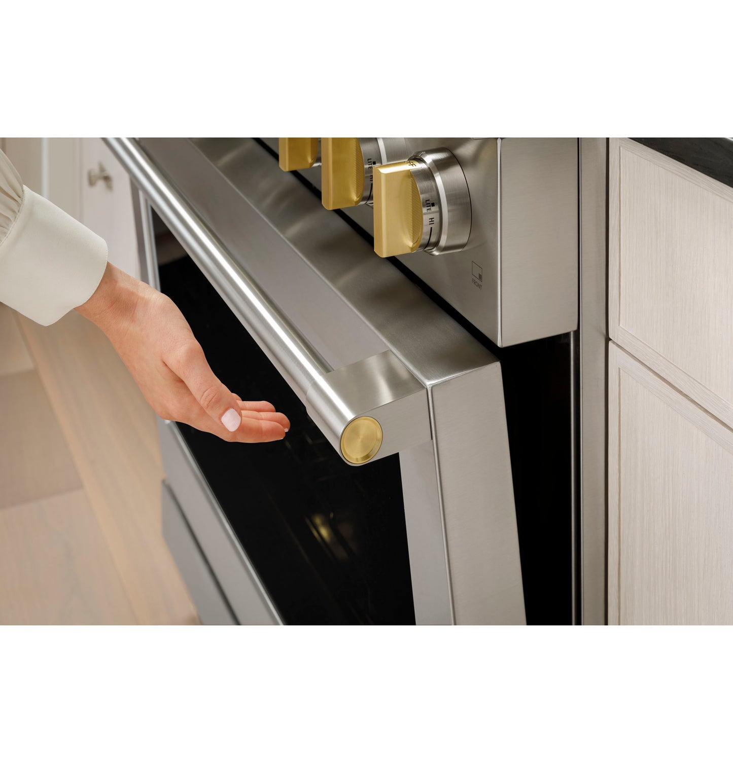 Hand reaching to open stainless steel oven door with brass-accented handle and control knobs in modern kitchen setting