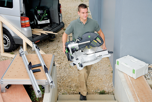 Man carrying Festool sliding compound miter saw up steps near workbench and van in home improvement setting