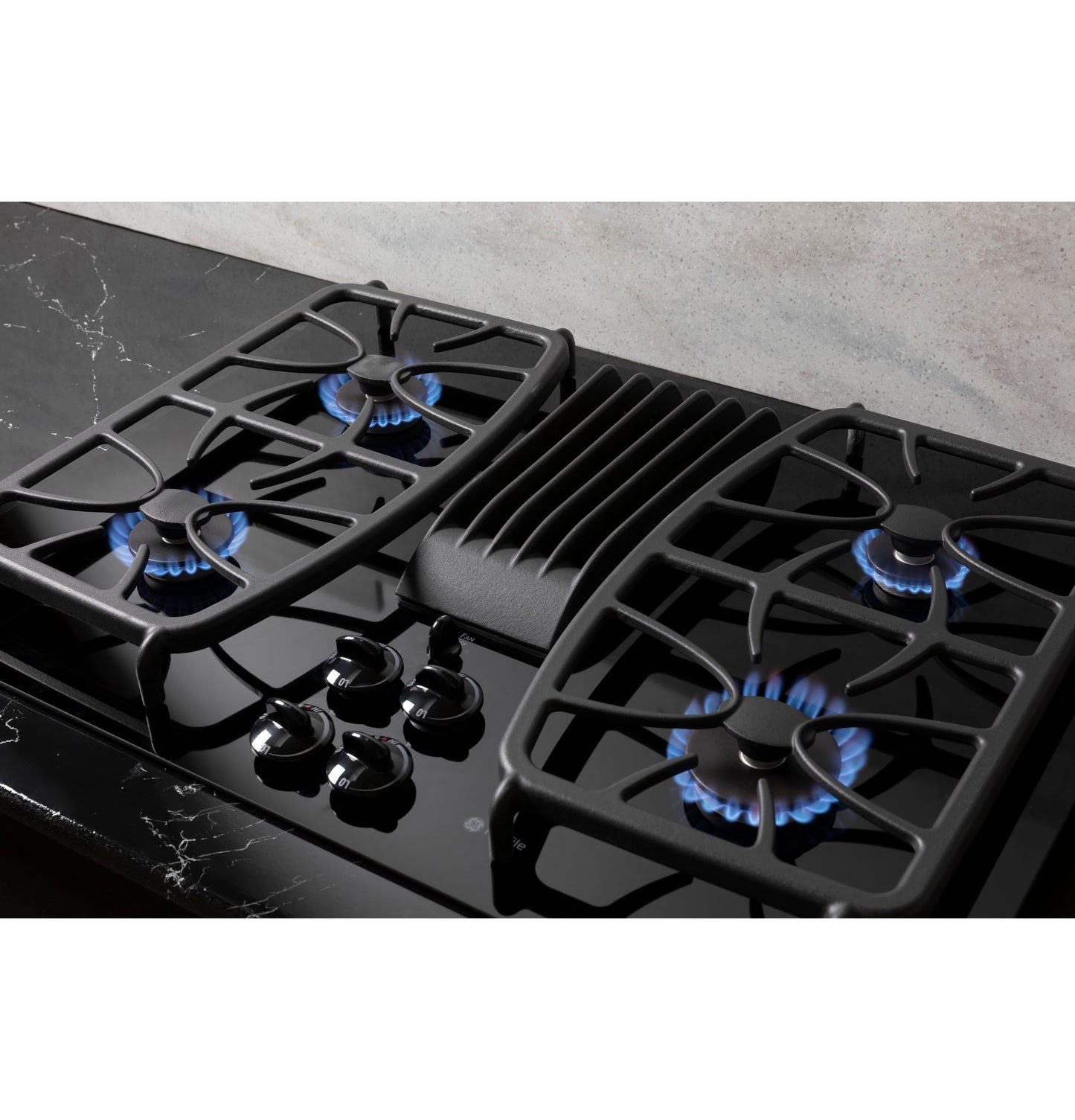 Black built-in gas cooktop with four burners lit and center downdraft vent in modern kitchen setting