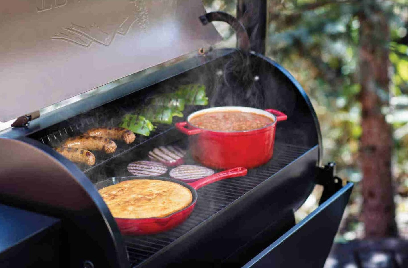 Bronze wood pellet grill cooking cast iron skillets and sausages with smoke rising in backyard setting
