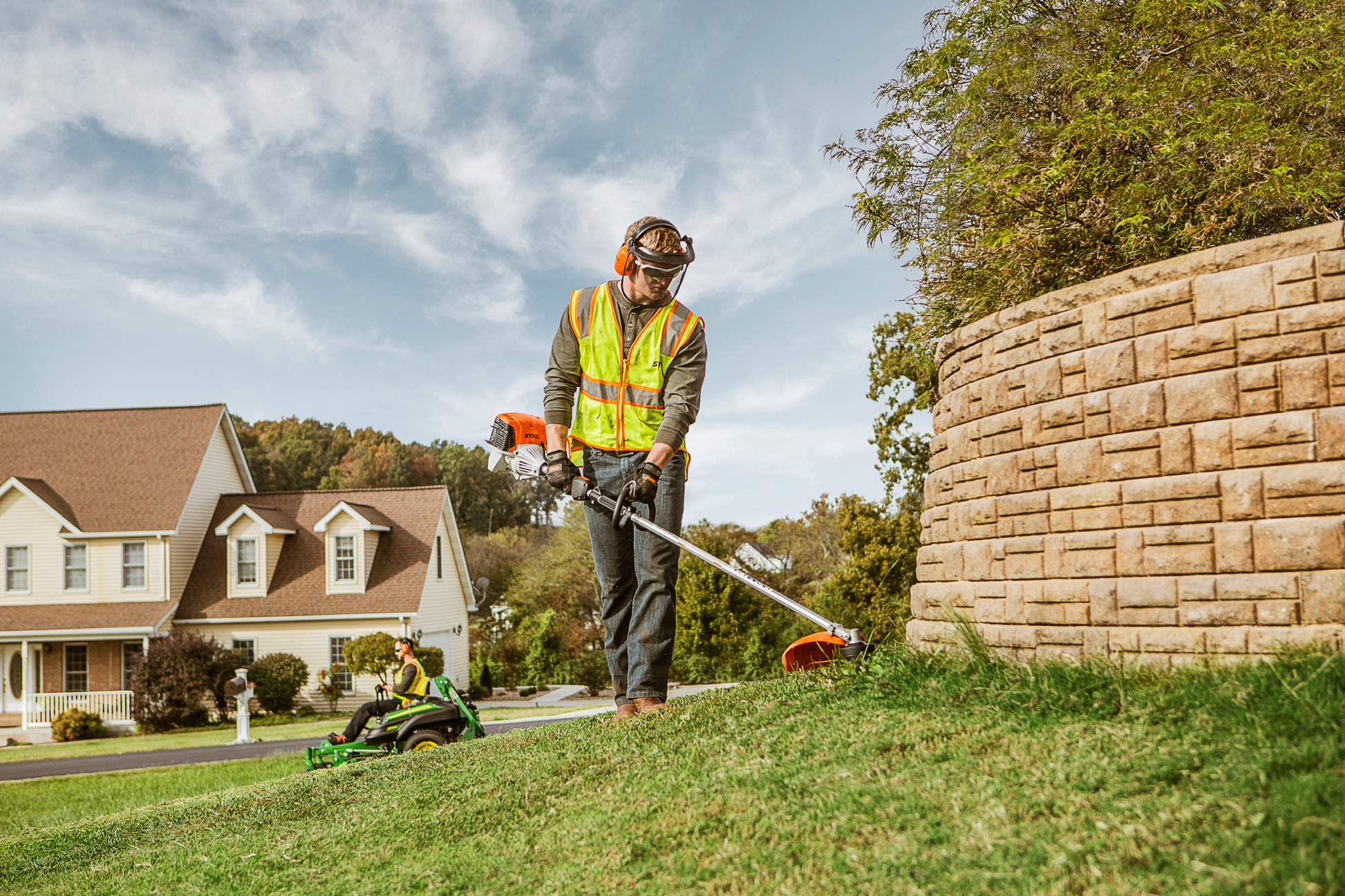 Man wearing safety gear using a straight shaft trimmer to edge grass near a stone retaining wall in home improvement setting