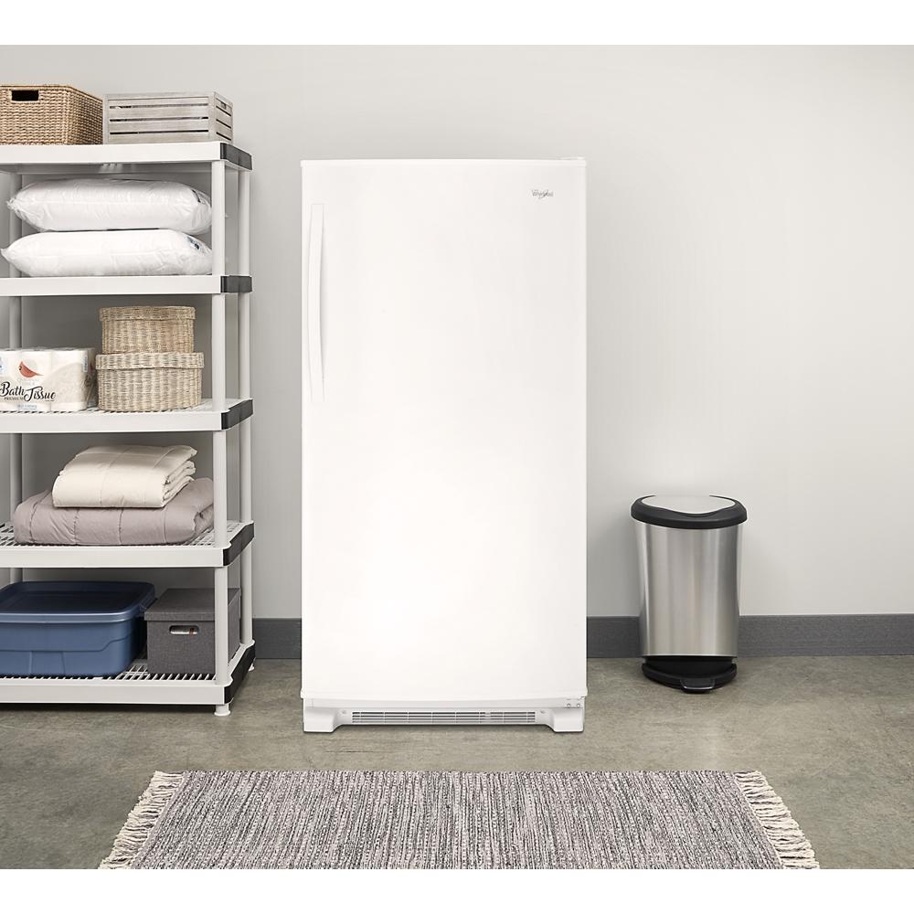 White upright freezer with bottom ventilation grille in a minimalist laundry room setting with shelving and stainless steel trash can