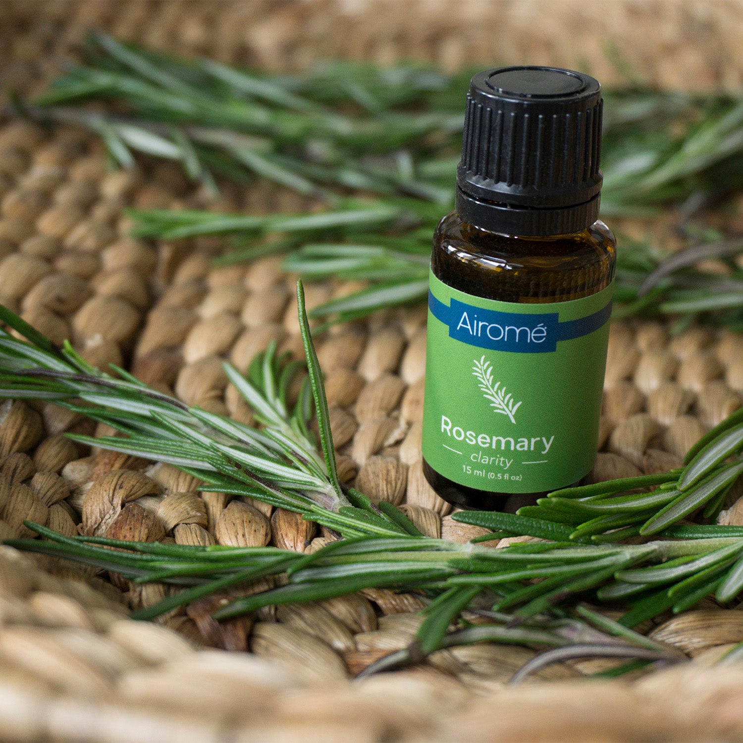 Small amber glass bottle of rosemary essential oil surrounded by fresh rosemary sprigs on woven decor surface
