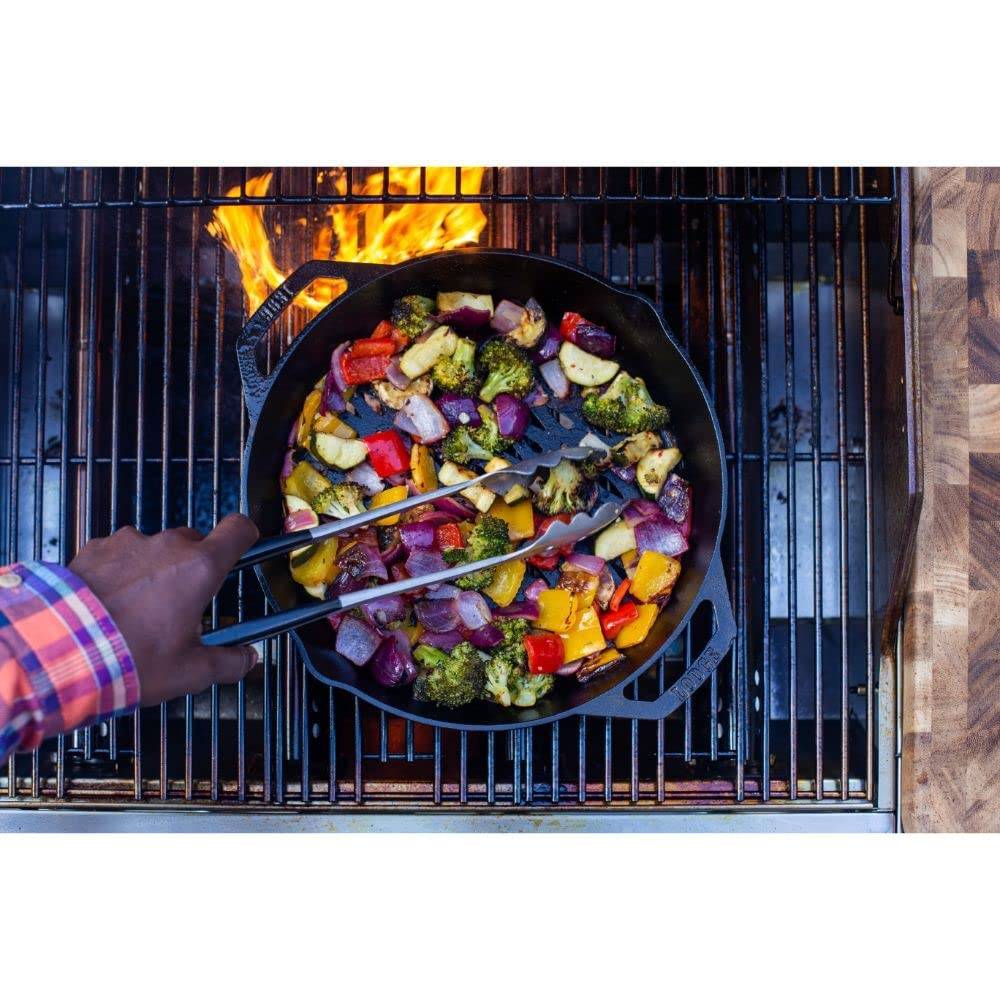Hand using tongs to cook mixed vegetables in a seasoned cast iron dual handle skillet over open flame grill in kitchenware collection