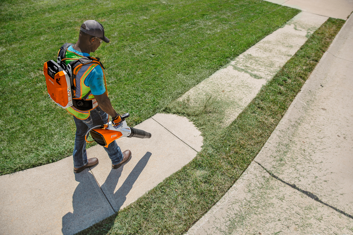 Man wearing safety vest using orange backpack-style battery blower to clear debris from sidewalk edge in home improvement task