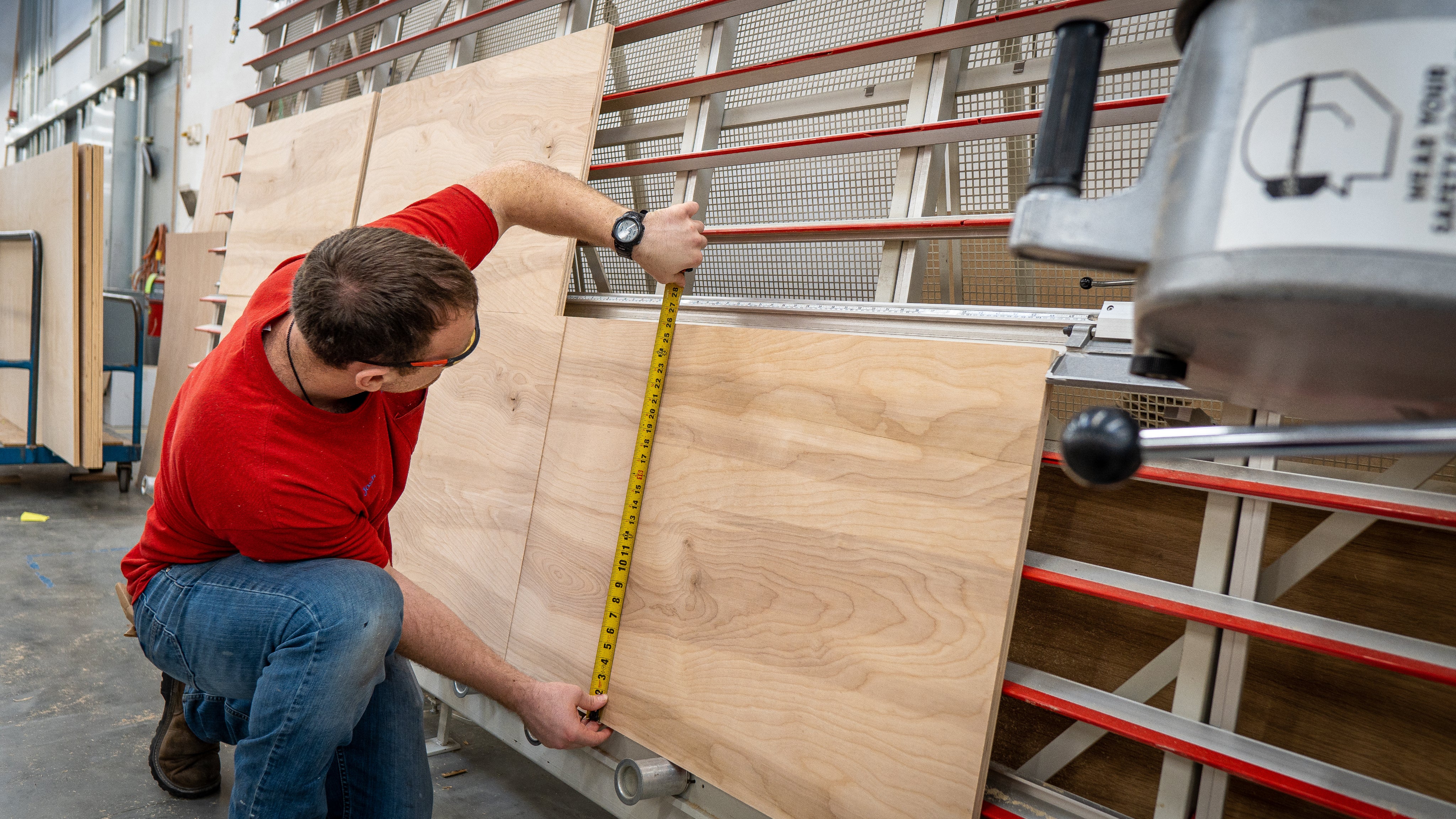 Man wearing a red shirt and jeans measuring a large wooden board with a tape measure in a workshop environment with metal racks.
