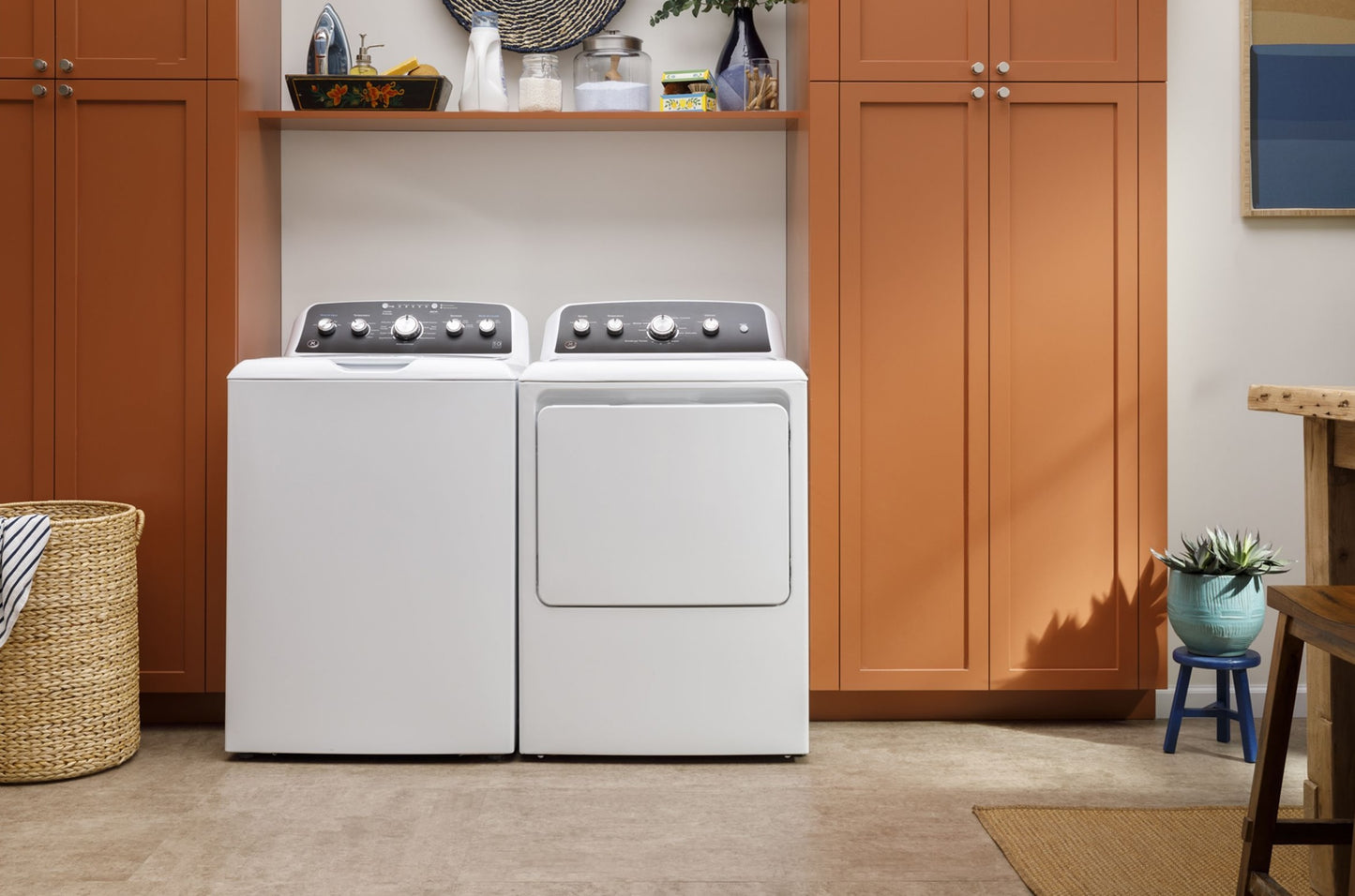 White top load washer and matching dryer set against orange cabinetry in a modern laundry room setting
