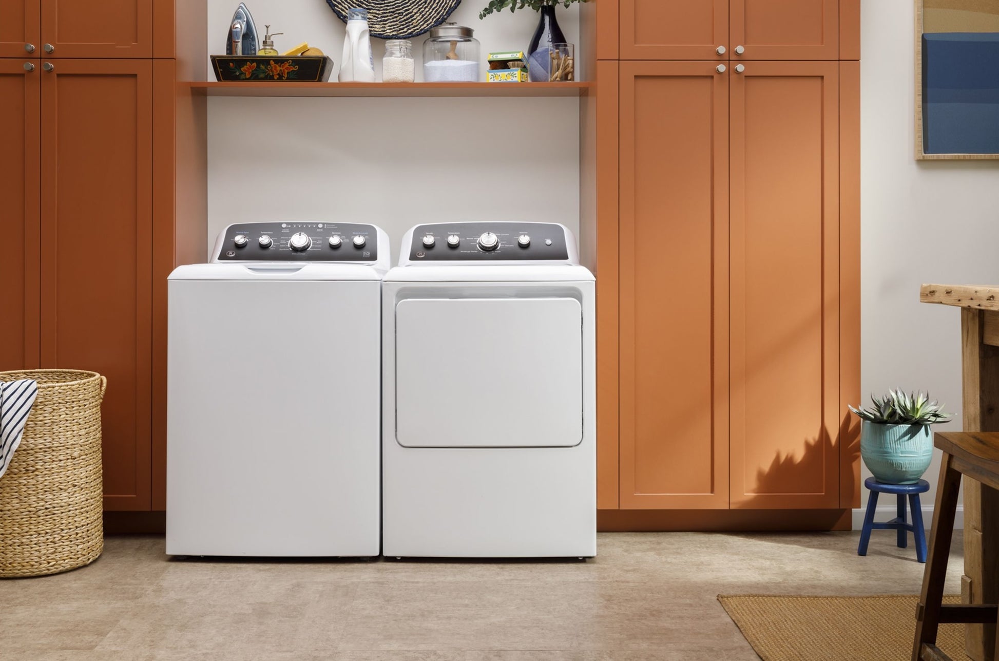 White top load washer and matching dryer set against orange cabinetry in a modern laundry room setting