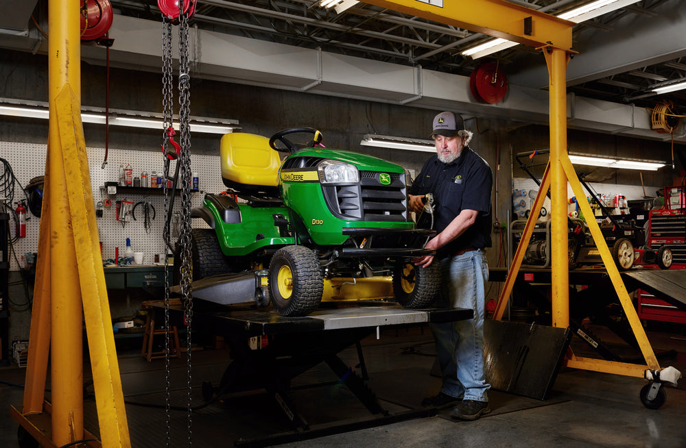 Man in a workshop adjusting a green and yellow lawn tractor elevated on a hydraulic lift surrounded by industrial tools and equipment.