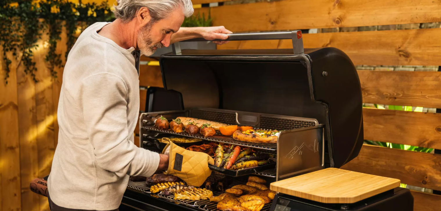 Man grilling assorted vegetables and meats on a black pellet grill with wooden side shelves in outdoor setting