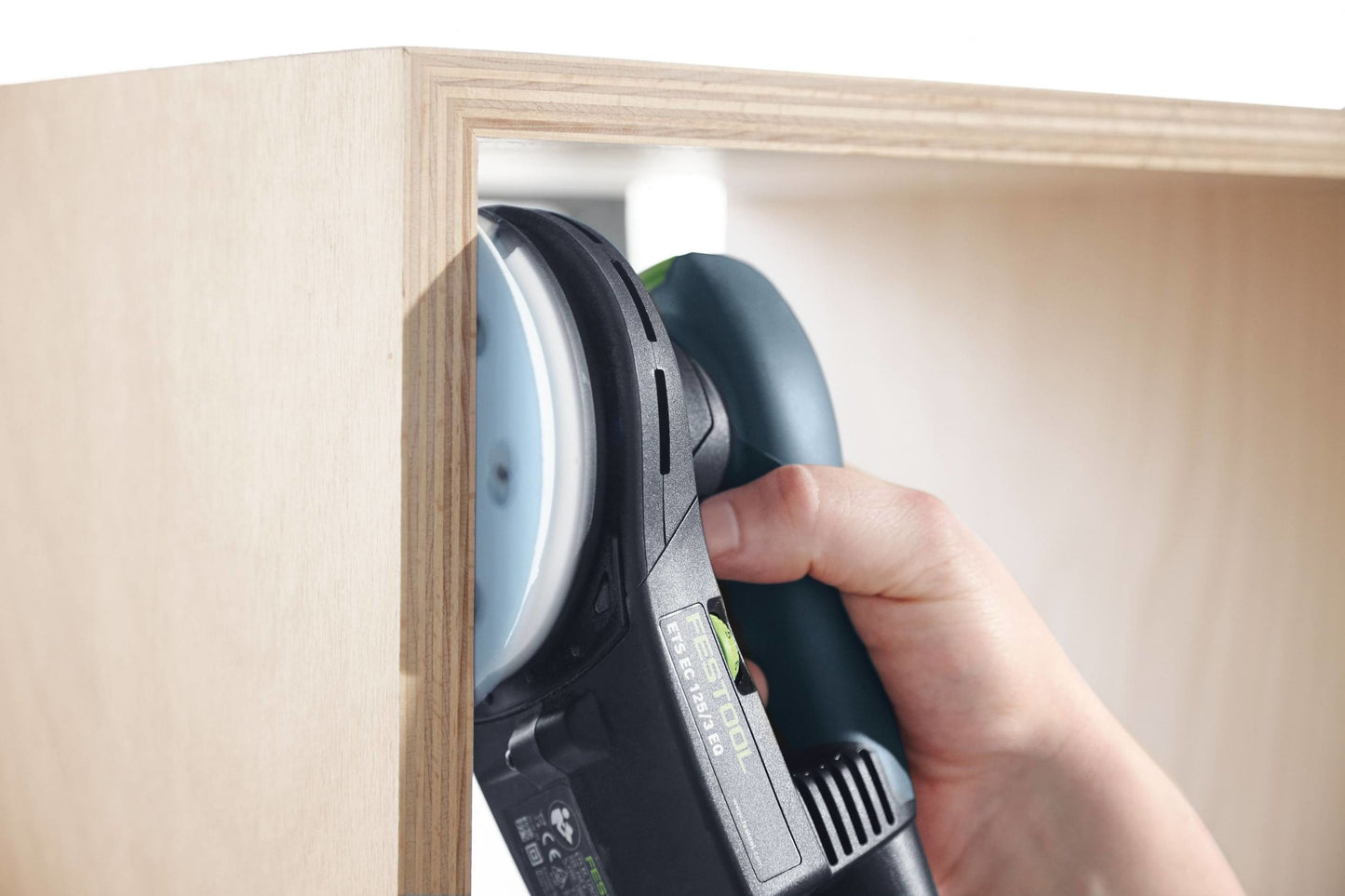 Hand gripping a Festool cordless random orbit sander smoothing the interior surface of a wood cabinet frame