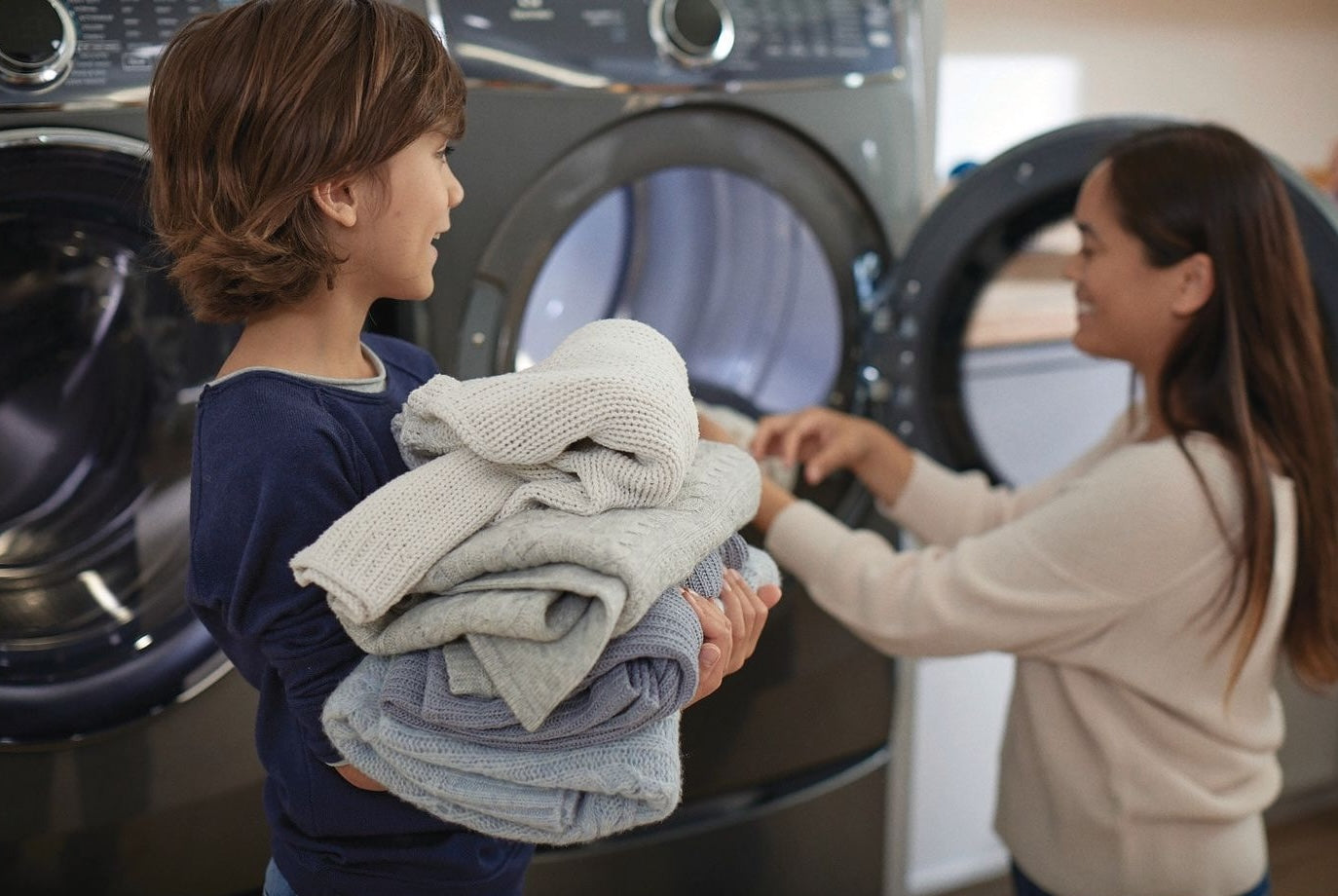 Child holding folded laundry while adult loads clothes into front load washer in modern laundry room
