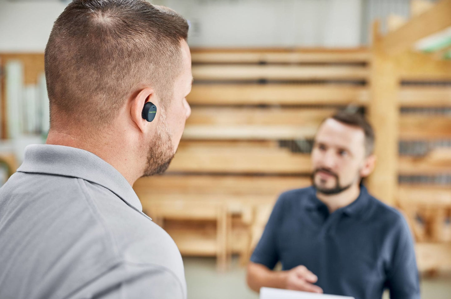 Man wearing black Bluetooth hearing protection earbuds in workshop while talking to coworker for safety and tools use