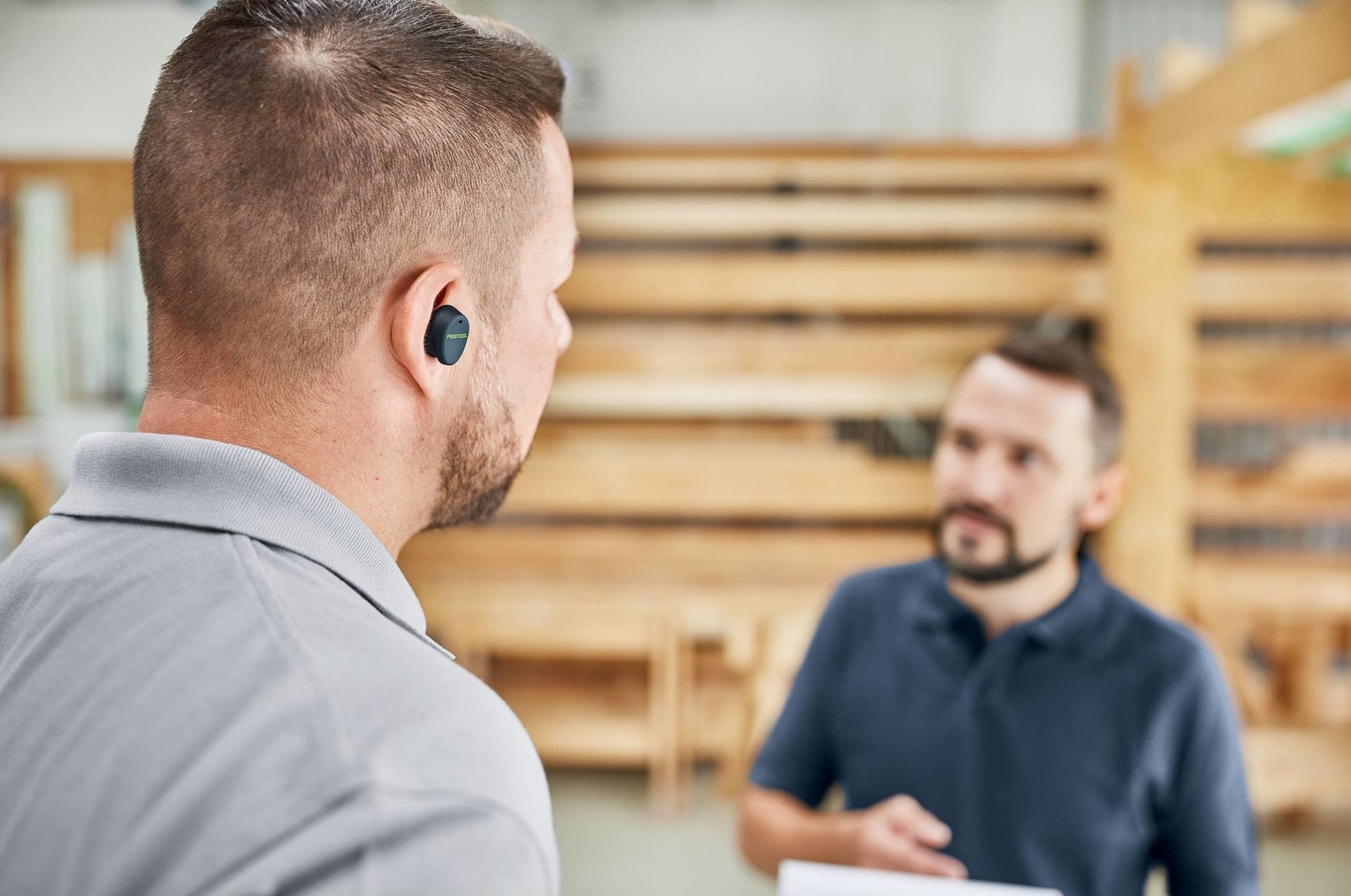 Man wearing black Bluetooth hearing protection earbuds in workshop while talking to coworker for safety and tools use