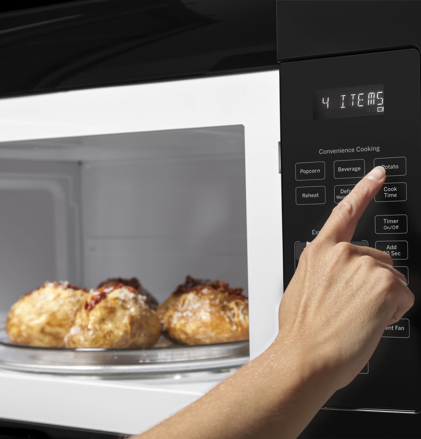 Hand selecting preset cooking option on black digital control panel of over-the-range microwave with baked potatoes inside