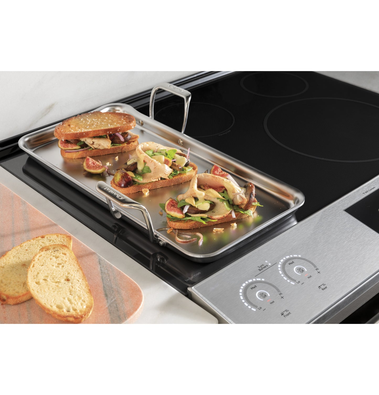 Stainless steel tray with sandwiches and figs on induction cooktop surface with digital front controls in kitchen setting