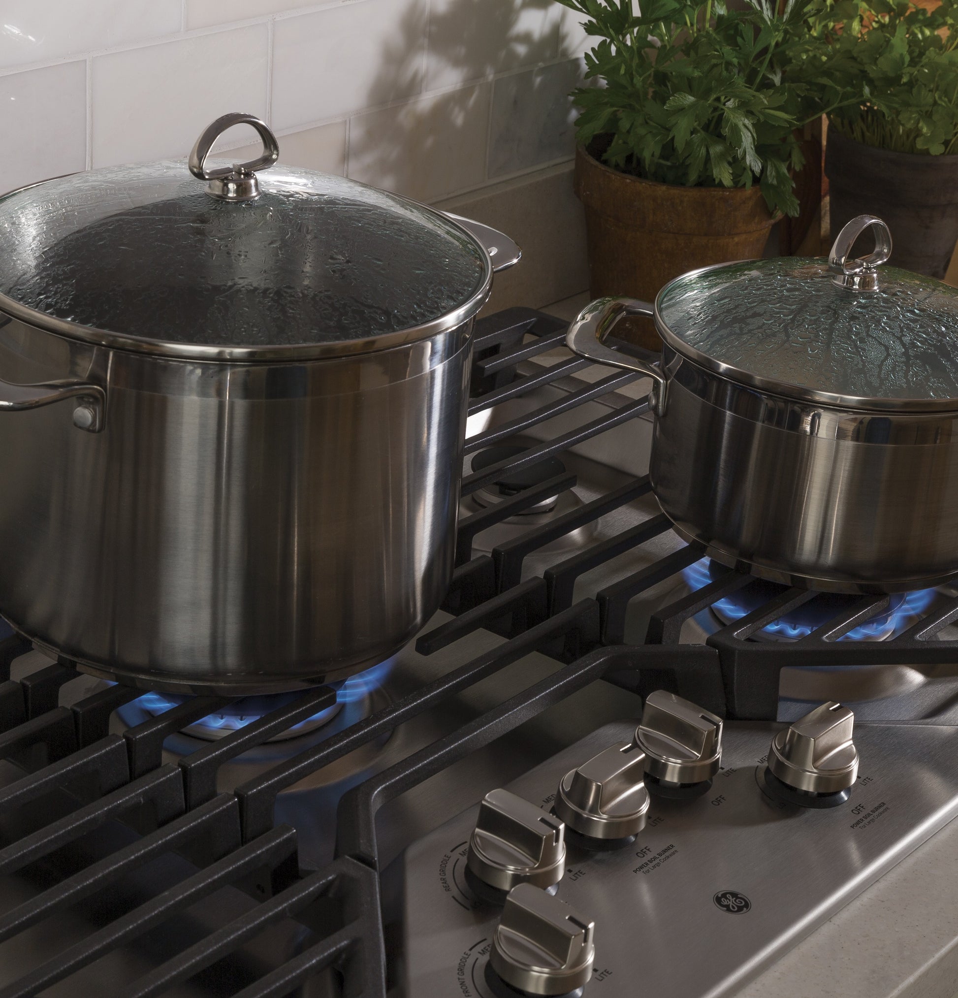 Two stainless steel pots with glass lids simmering on a built-in gas cooktop with blue flames and control knobs visible