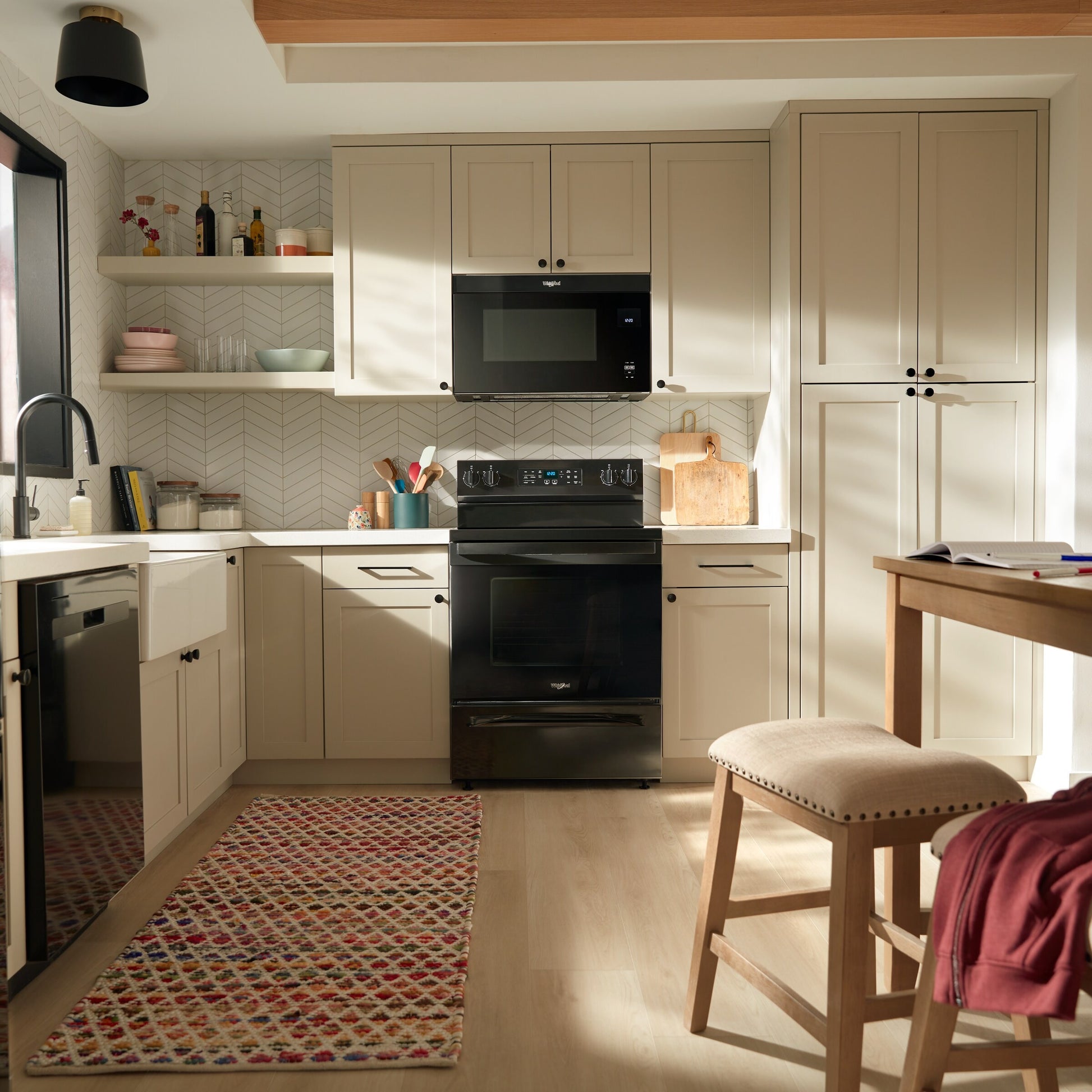 Modern kitchen with black electric range and microwave, cream cabinetry, wooden stool, and patterned rug in natural light