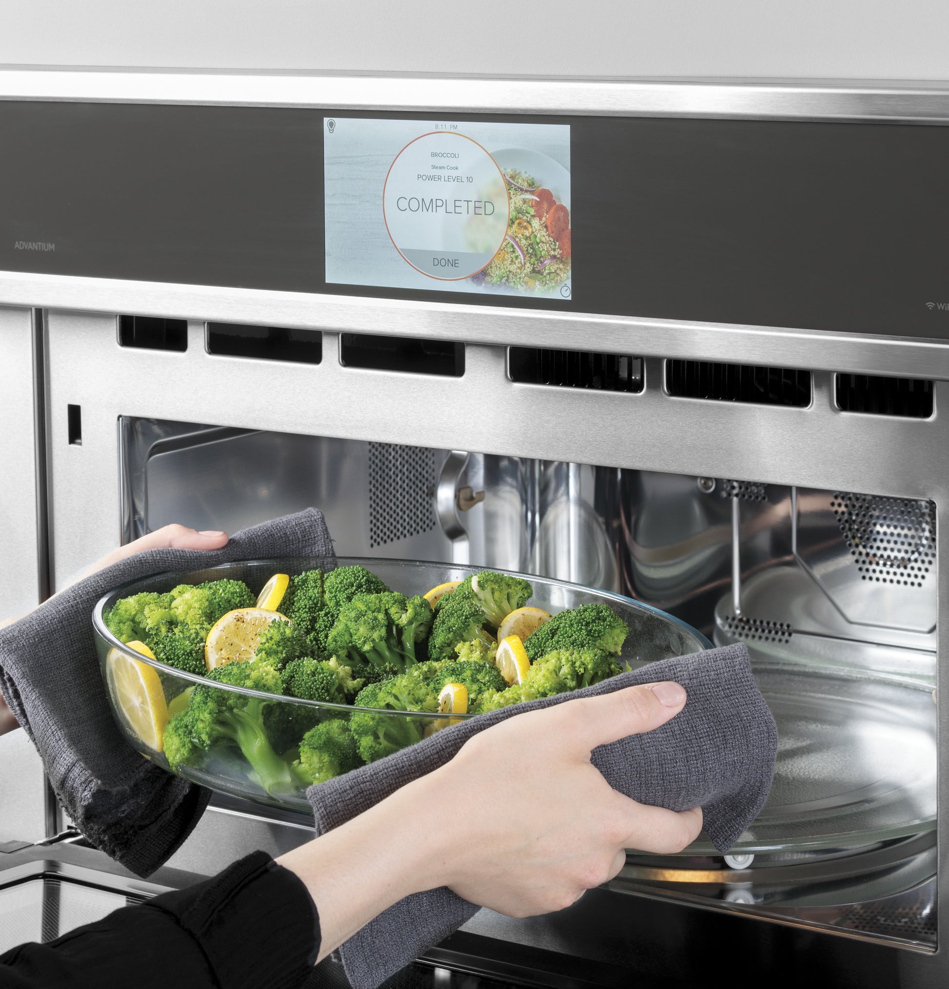 Hands removing a glass dish with steamed broccoli and lemon wedges from a stainless steel wall oven with digital display