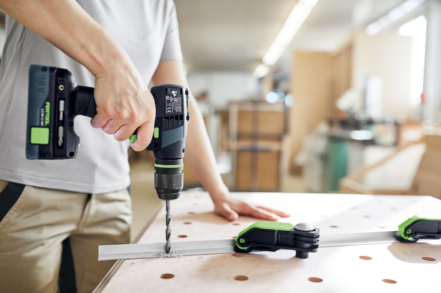 Cordless drill with green accents drilling into wood, held by person in woodworking shop surrounded by tools and clamps
