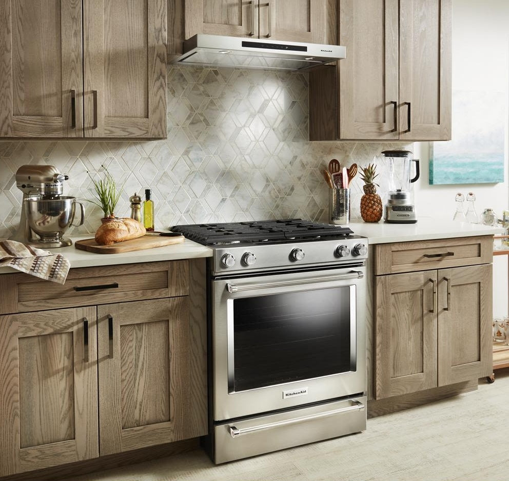 Stainless steel range with under-cabinet vent hood in kitchen featuring wood cabinetry, backsplash, and countertop appliances