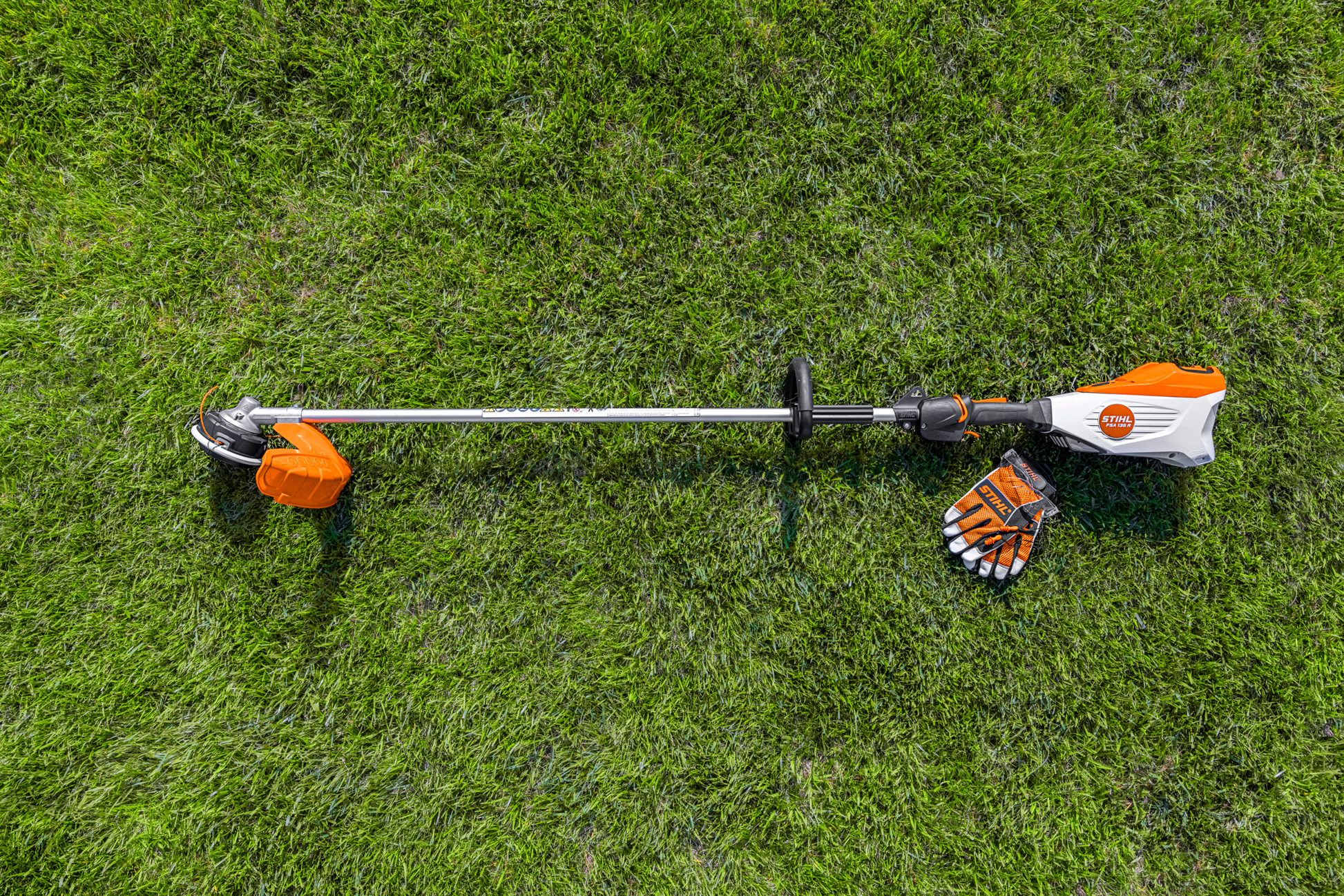 Orange and white battery-powered trimmer with metal shaft and gloves on green grass for home improvement tasks