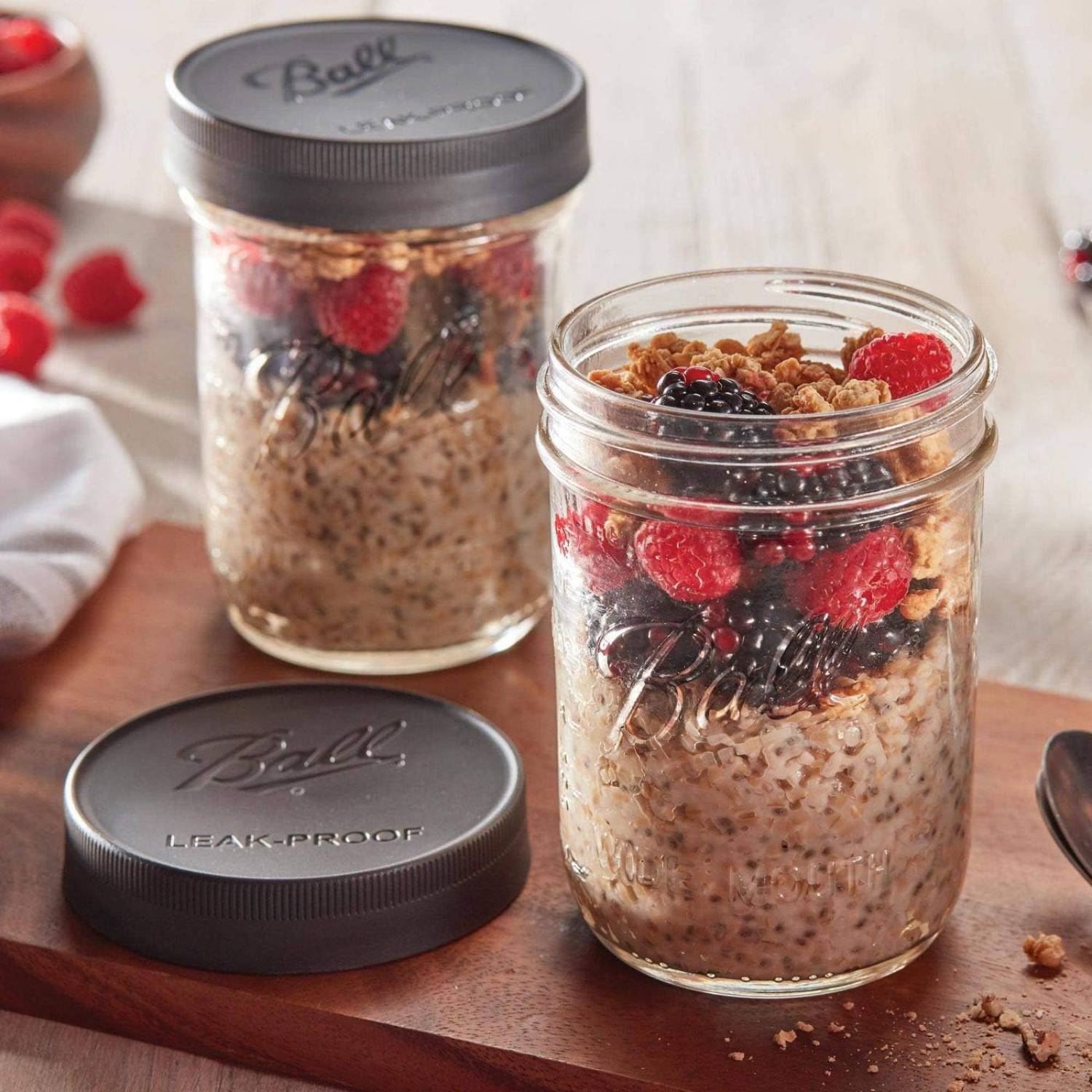 Two clear wide-mouth jars with black leak-proof lids filled with granola and fresh berries on a kitchenware surface