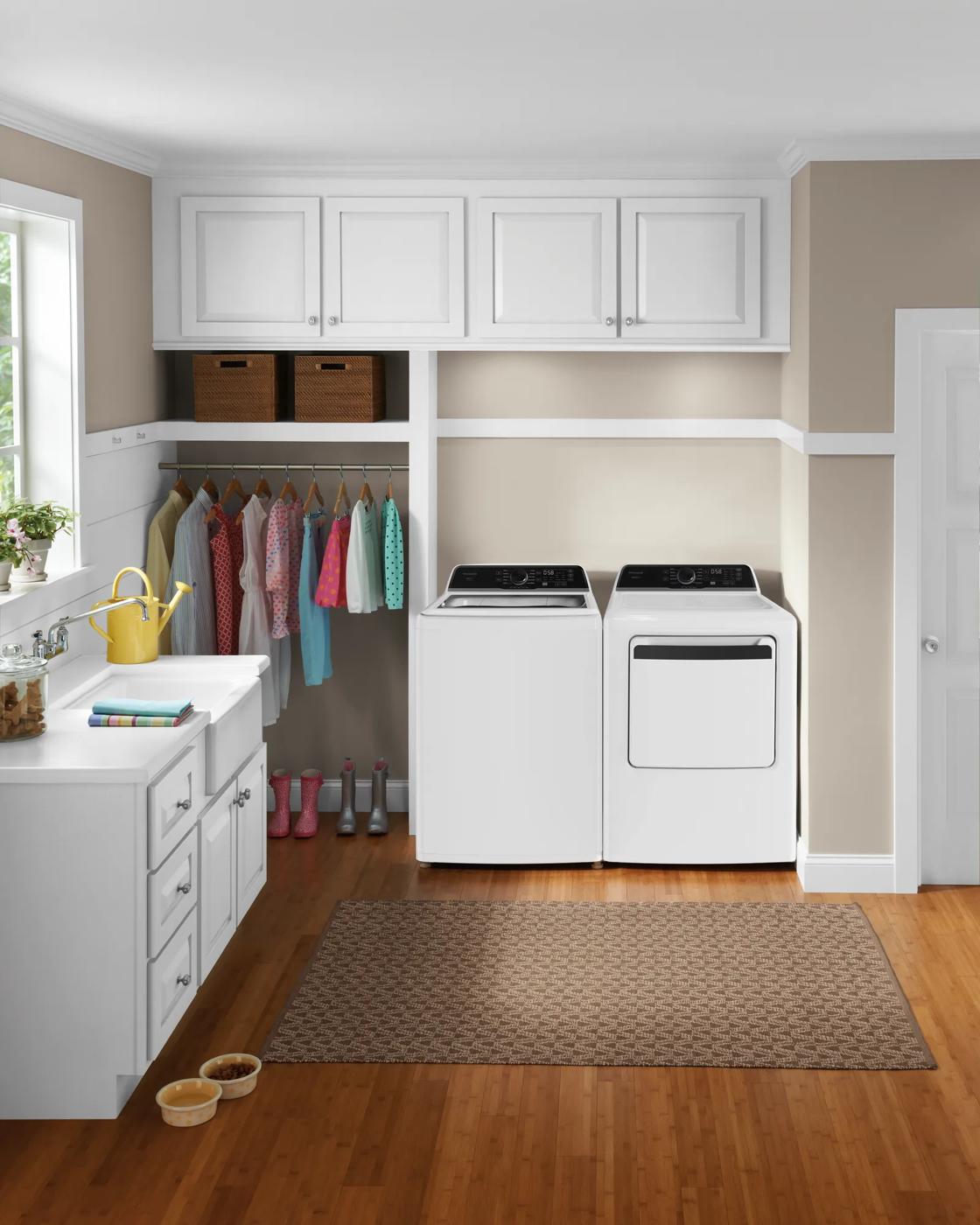 Bright laundry room with white washer and dryer set beneath white cabinets, adjacent to organized storage and hardwood floor rug