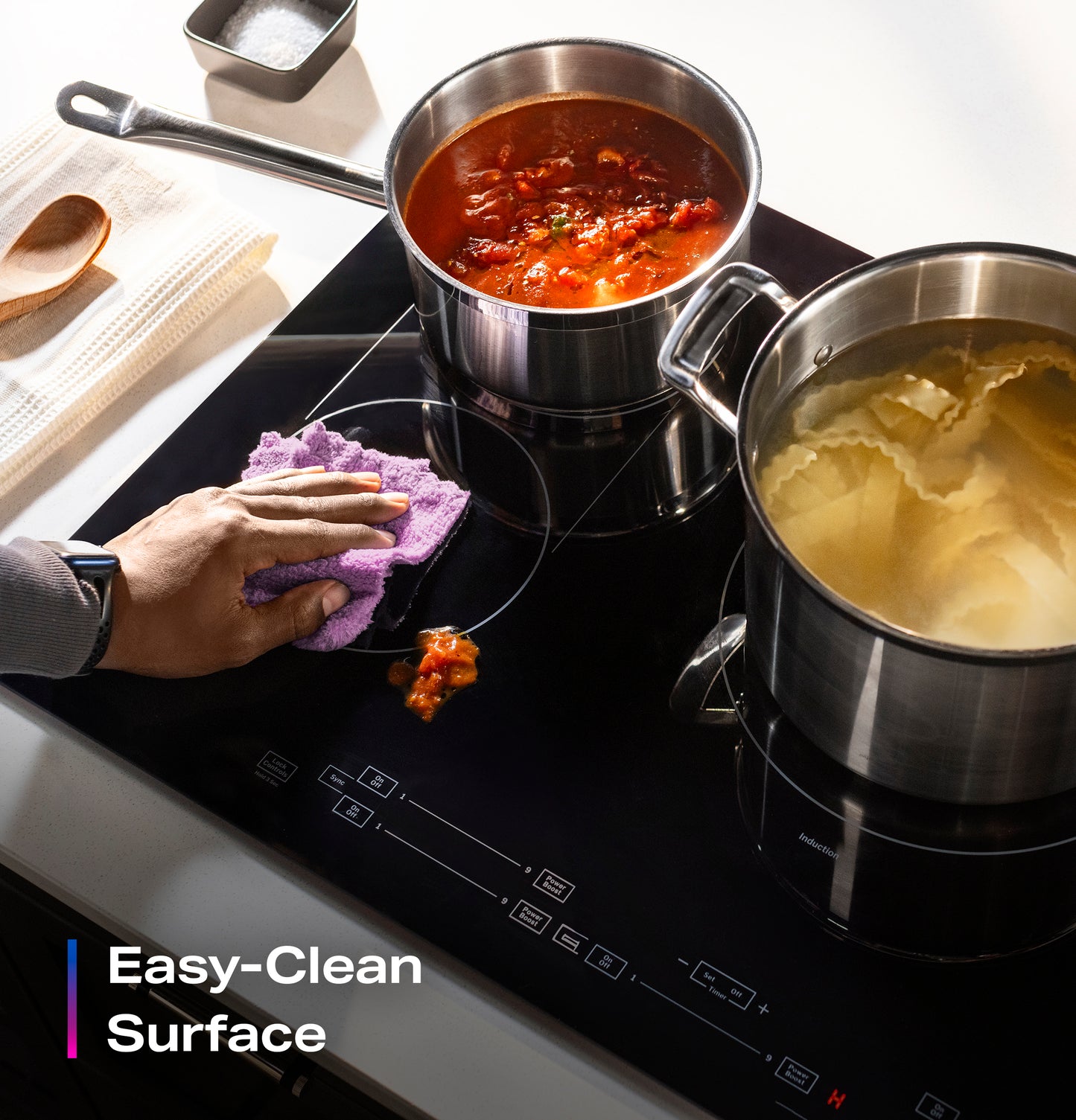 Hand wiping spill on black built-in induction cooktop with two stainless steel pots cooking sauce and pasta