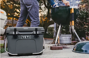 Person standing beside a dark green charcoal grill with a lid resting nearby and a gray heavy-duty cooler on a concrete surface outdoors at dusk.