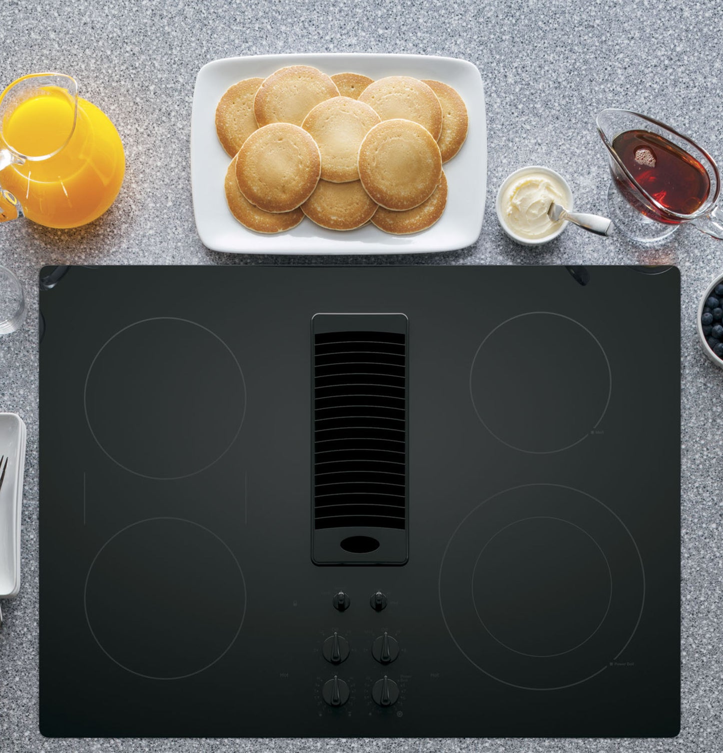 Black downdraft electric cooktop with four burners and front knobs surrounded by breakfast items from cooktops collection