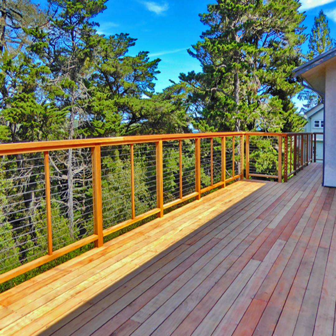 Outdoor wooden deck featuring cable railing system with wood posts surrounded by lush green trees under blue sky