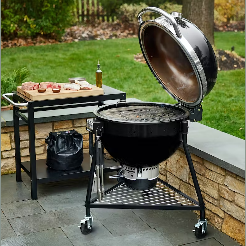 Black charcoal grill with open lid next to prep table holding raw meats and olive oil for outdoor cooking and grilling