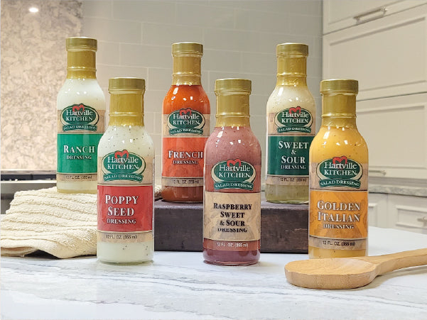 Bottles of Hartville Kitchen salad dressings in various flavors arranged on a kitchen countertop with a wooden spoon and towel nearby.