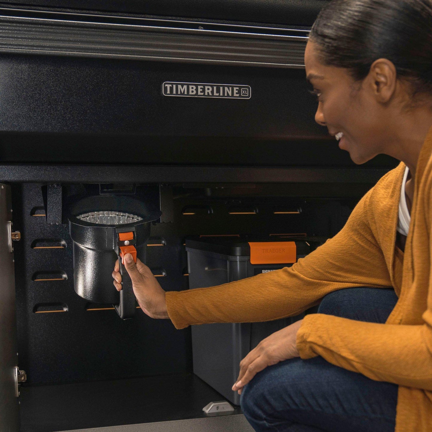 Woman removing grease and ash keg liner from black Timberline grill for outdoor living and grill accessories under 50 collection