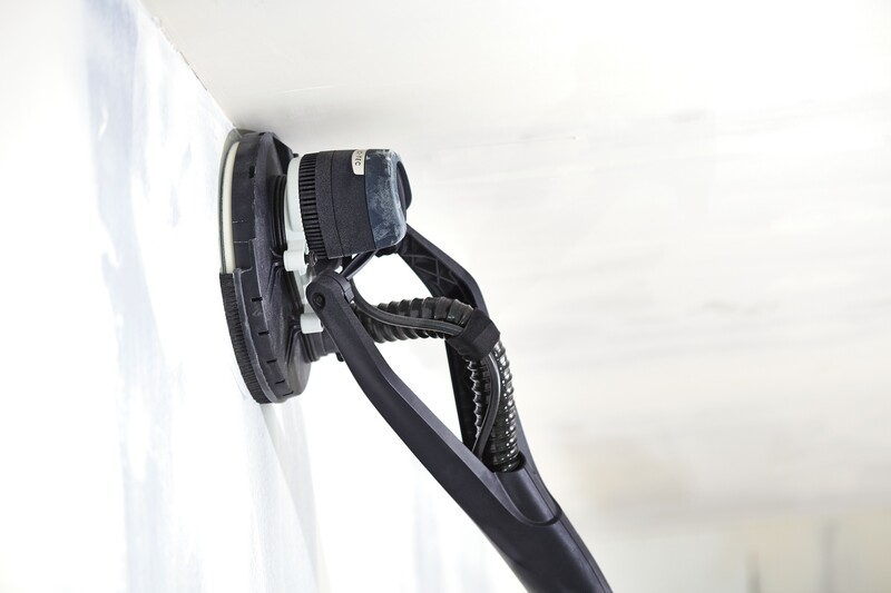 Black handheld drywall sander with ergonomic handle and circular sanding pad smoothing a white ceiling surface