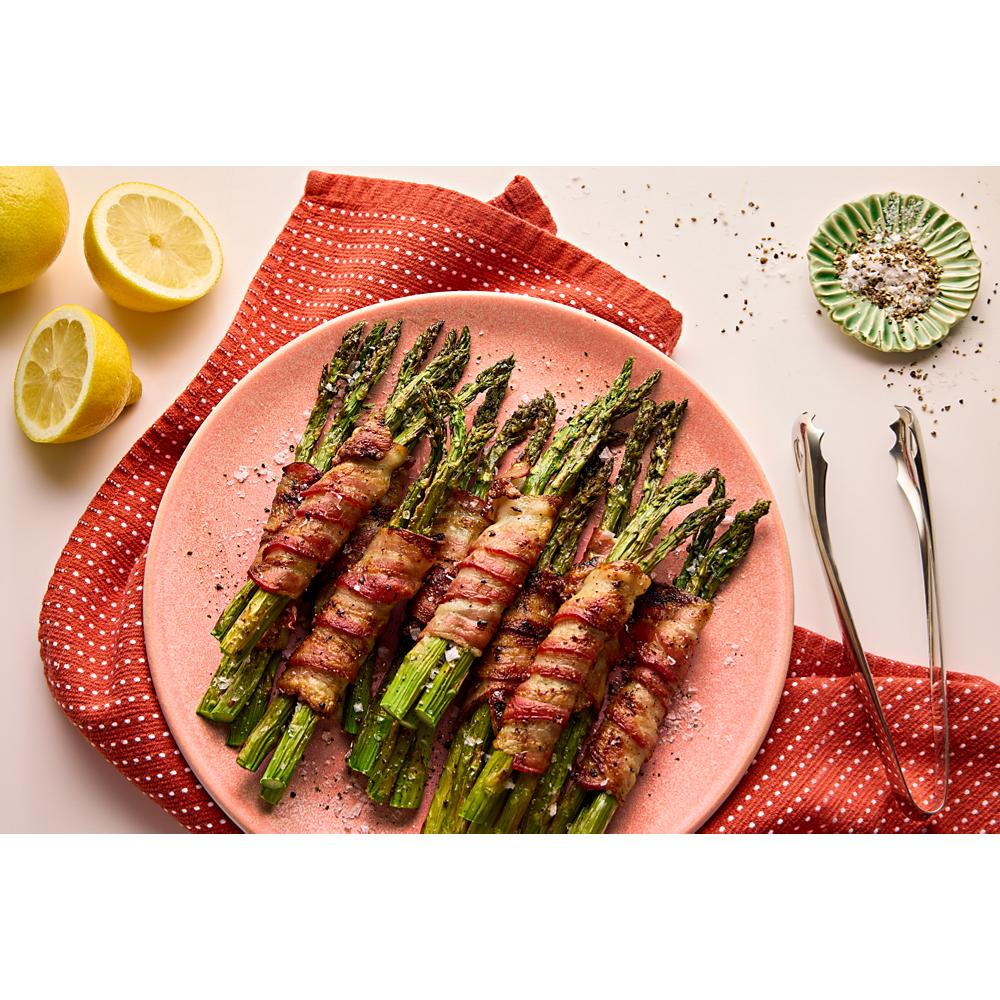 Bacon-wrapped asparagus bundles on pink plate over red cloth, with lemon halves, seasoning dish, and metal tongs nearby
