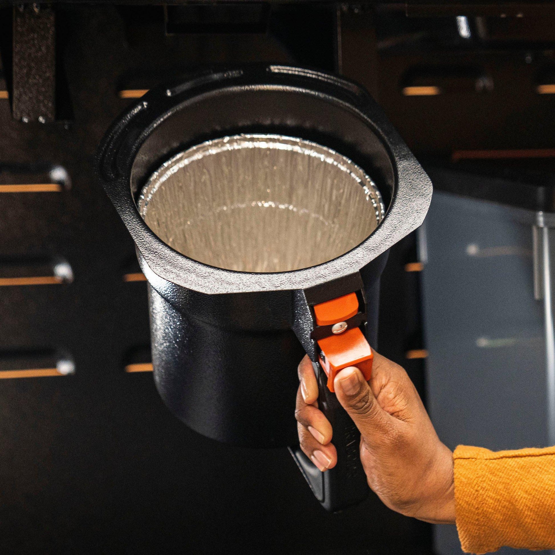 Hand holding black grease and ash keg with disposable aluminum liner for grill cleaning and outdoor living tools